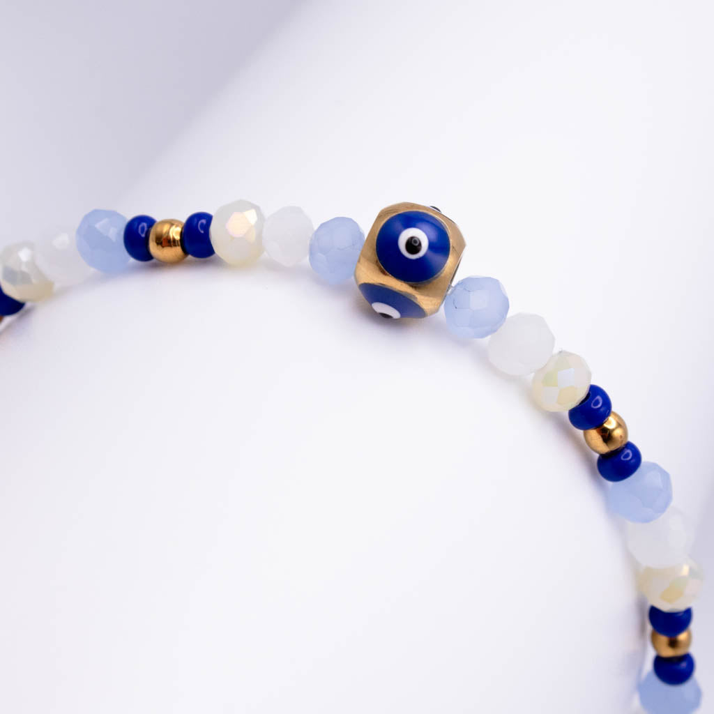 A beaded bracelet featuring light blue and milky white faceted beads, dark blue evil eye beads, and small gold spacer beads, with a prominent gold cube charm displaying a blue evil eye bead.