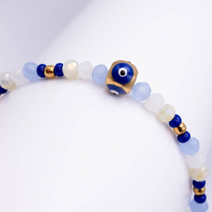 A beaded bracelet featuring light blue and milky white faceted beads, dark blue evil eye beads, and small gold spacer beads, with a prominent gold cube charm displaying a blue evil eye bead.