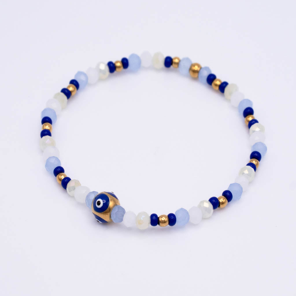 A beaded stretch bracelet featuring light blue, white, and dark blue beads, accented with small gold spacer beads and a central blue and white evil eye charm, shown on a plain white background.