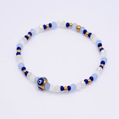 A beaded stretch bracelet featuring light blue, white, and dark blue beads, accented with small gold spacer beads and a central blue and white evil eye charm, shown on a plain white background.