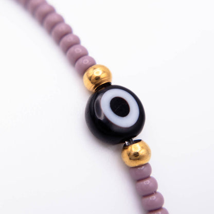 A close-up of a beaded necklace featuring a central black and white evil eye bead, flanked by shiny gold spacer beads, with lavender-purple round beads on either side, set against a clean white background.