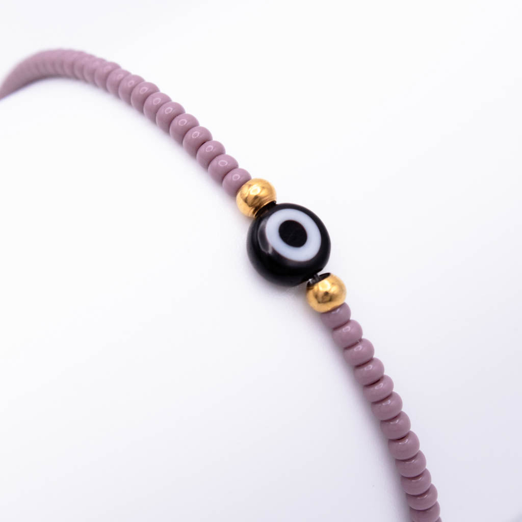 A beaded bracelet featuring dusty purple round beads, a central black and white evil eye charm, and two small gold spacer beads, shown on a plain white background.