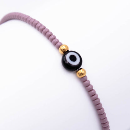 A beaded bracelet featuring dusty purple round beads, a central black and white evil eye charm, and two small gold spacer beads, shown on a plain white background.