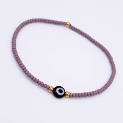 A beaded stretch bracelet featuring a dusty purple bead strand, a central black evil eye charm with white details, and small gold spacer beads, shown on a plain white background.