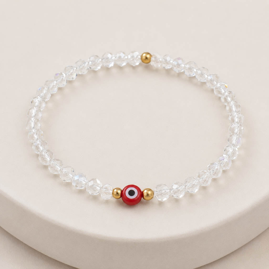 A beaded stretch bracelet made of clear faceted beads, featuring a central red evil eye charm with white, black, and gold details, flanked by two small gold spacer beads, displayed on a light beige surface.