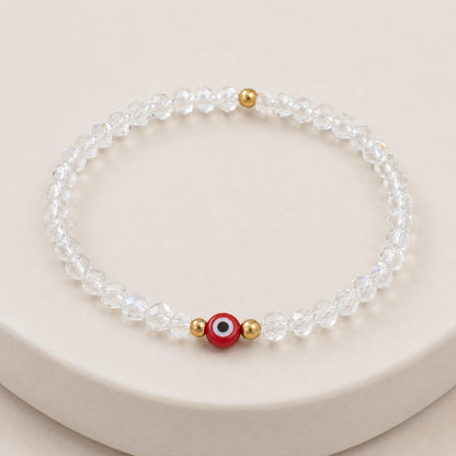A beaded stretch bracelet made of clear faceted beads, featuring a central red evil eye charm with white, black, and gold details, flanked by two small gold spacer beads, displayed on a light beige surface.