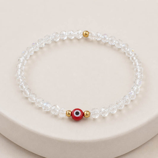 A beaded stretch bracelet made of clear faceted beads, featuring a central red evil eye charm with white, black, and gold details, flanked by two small gold spacer beads, displayed on a light beige surface.