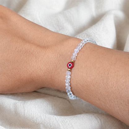 A clear faceted bead bracelet with a central red evil eye charm, shown worn on a person's wrist against a soft beige fabric background.