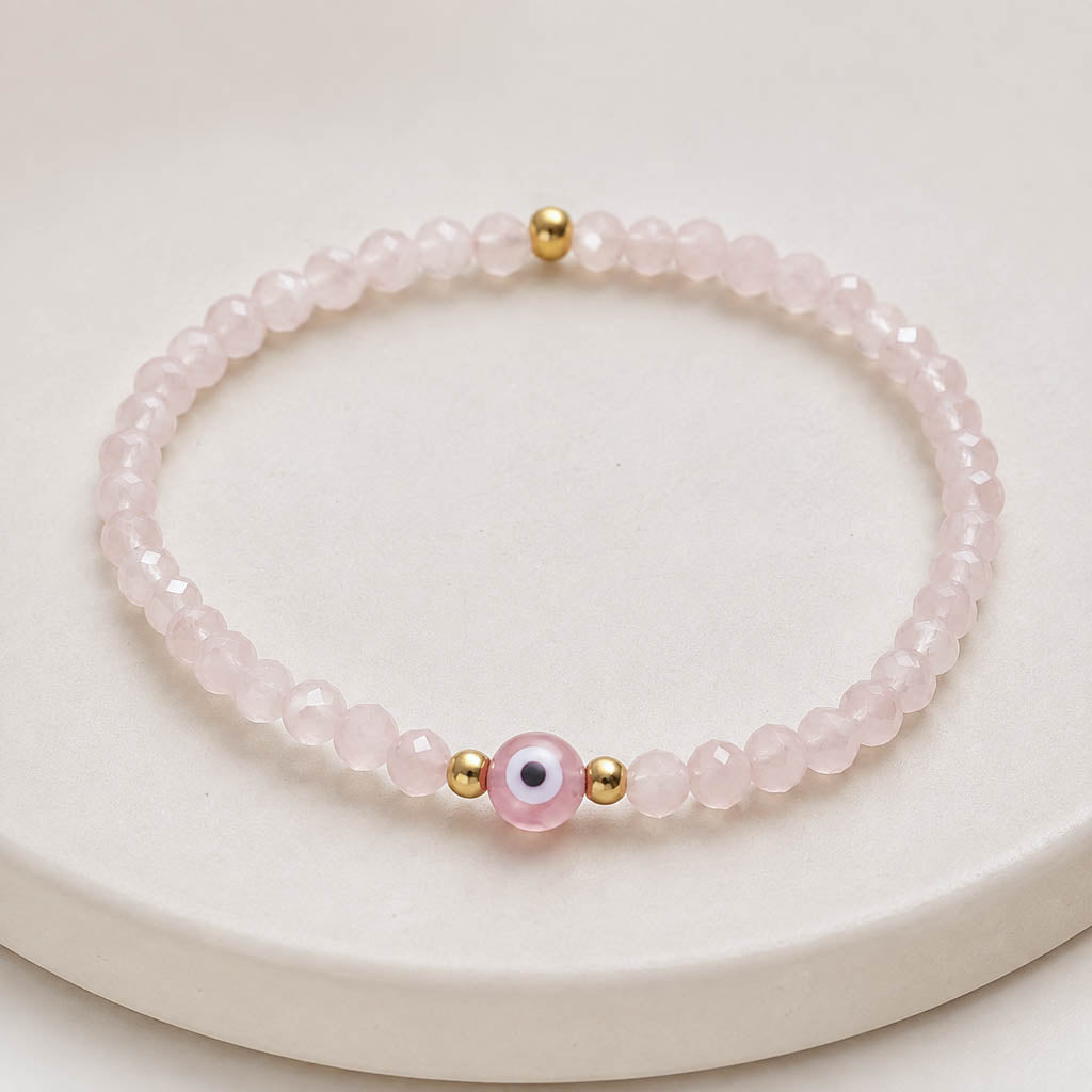 A pink beaded bracelet with a central evil eye charm and small gold spacer beads, displayed on a round cream-colored surface.