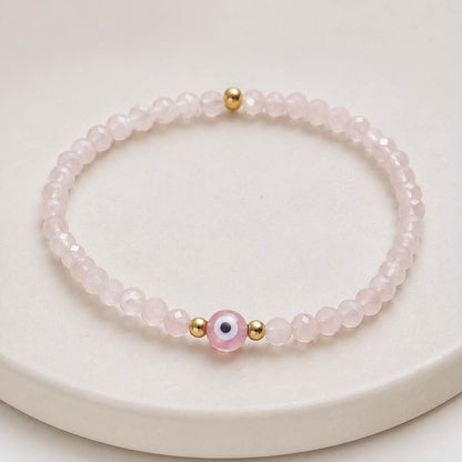 A pink beaded bracelet with a central evil eye charm and small gold spacer beads, displayed on a round cream-colored surface.