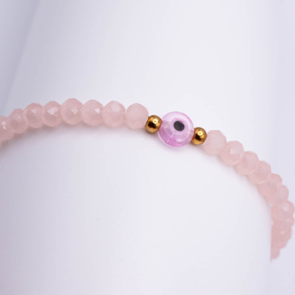 A pink beaded bracelet with faceted round beads and a central pink evil eye charm with gold spacer beads, displayed on a white surface.
