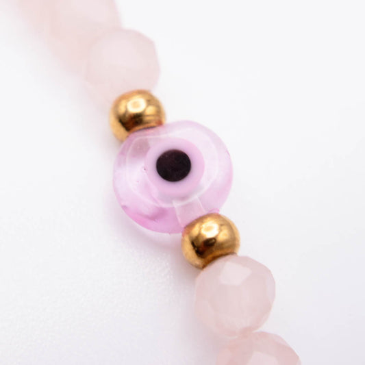 A close-up of a beaded strand featuring translucent light pink faceted beads, a central pink glass evil eye bead with a black pupil, and shiny gold spacer beads on a white background.