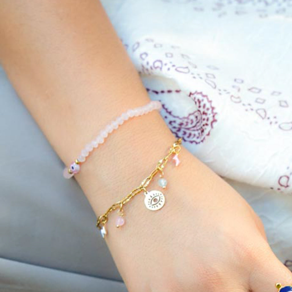 A close-up of a person's wrist wearing two delicate bracelets: a pale pink beaded bracelet and a gold-tone charm bracelet with a circular sun face charm and small pink beads.
