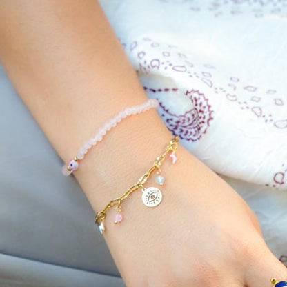 A close-up of a person's wrist wearing two delicate bracelets: a pale pink beaded bracelet and a gold-tone charm bracelet with a circular sun face charm and small pink beads.