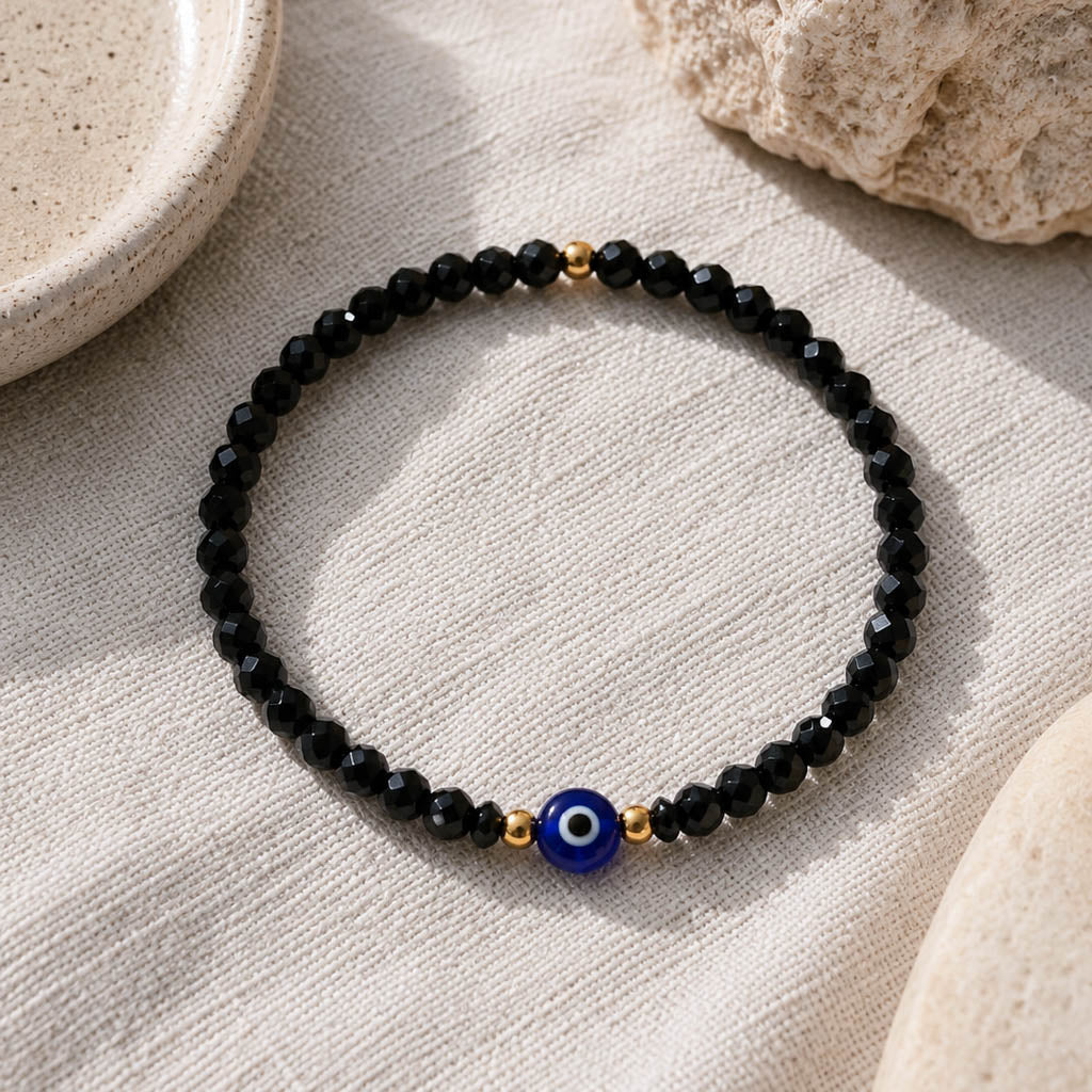 A beaded stretch bracelet featuring faceted black beads, small gold spacer beads, and a central blue evil eye charm, displayed on a textured beige linen surface with neutral stone props.