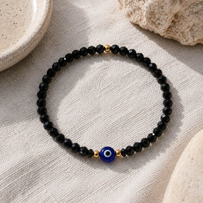 A beaded stretch bracelet featuring faceted black beads, small gold spacer beads, and a central blue evil eye charm, displayed on a textured beige linen surface with neutral stone props.