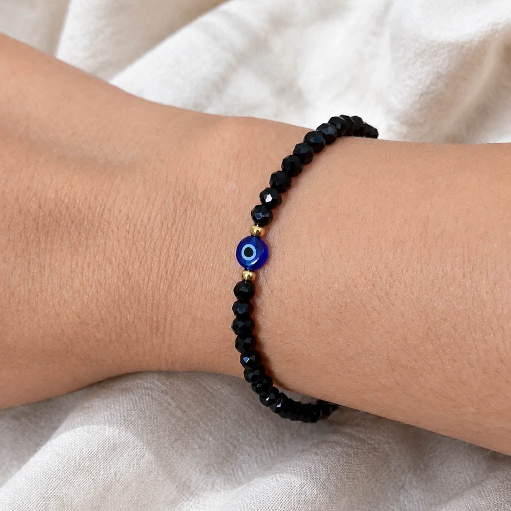 A black beaded bracelet with a central blue and white evil eye charm, shown worn on a person's wrist against a soft white fabric background.