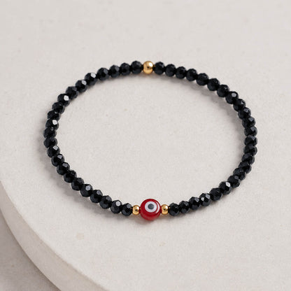 A beaded stretch bracelet featuring faceted black beads, small gold spacer beads, and a central red evil eye charm with white and black details, displayed on a light beige textured surface.