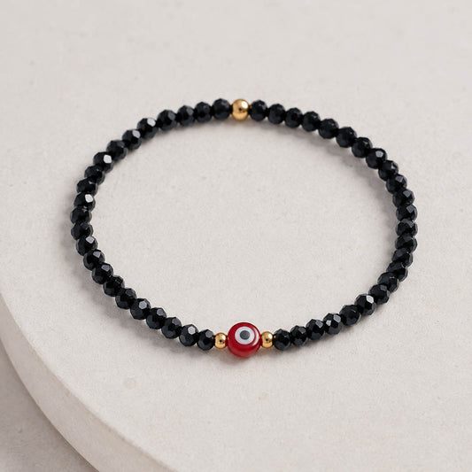 A beaded stretch bracelet featuring faceted black beads, small gold spacer beads, and a central red evil eye charm with white and black details, displayed on a light beige textured surface.