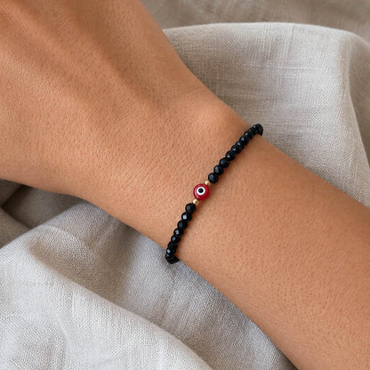 A black beaded bracelet with a red, white, and black evil eye charm worn on a person's wrist, set against a neutral beige linen background.