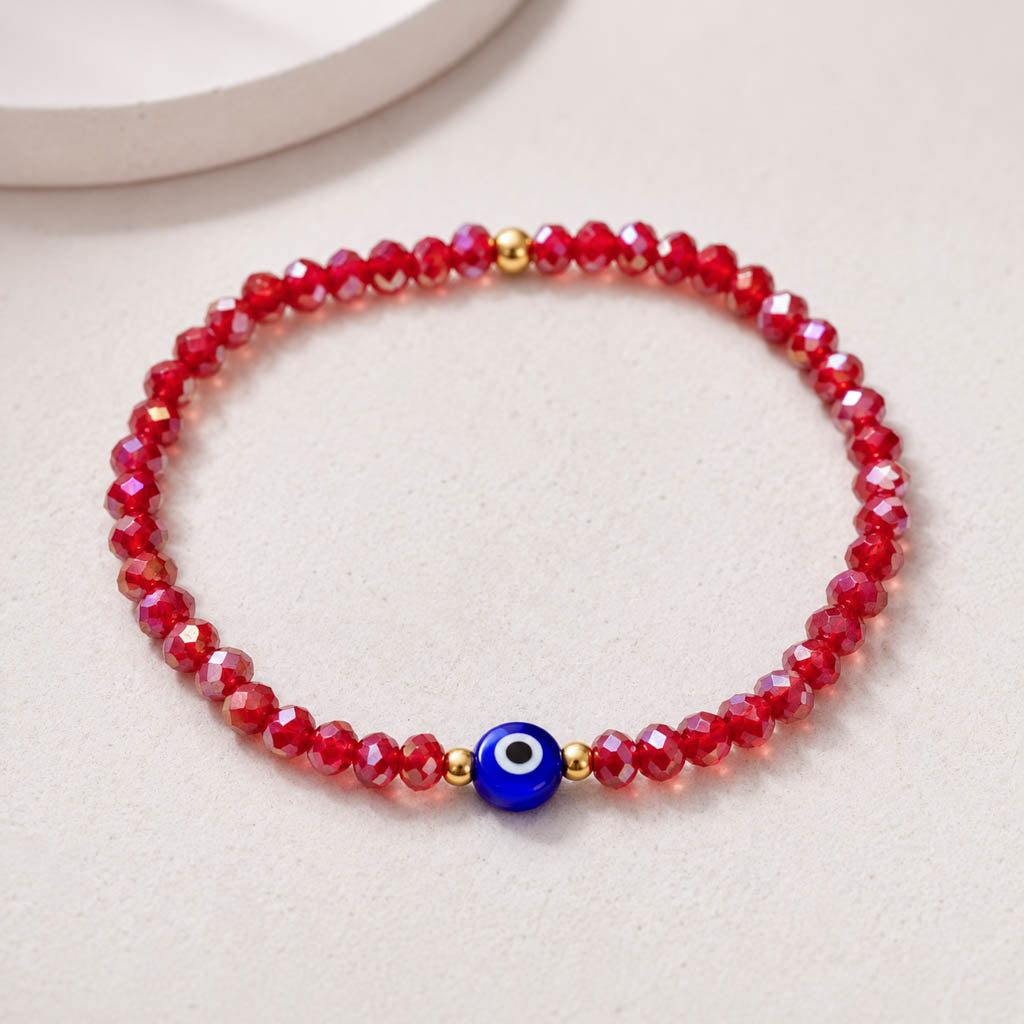 A beaded stretch bracelet made of faceted red beads with a central blue evil eye charm and small gold spacer beads, photographed on a light neutral surface.