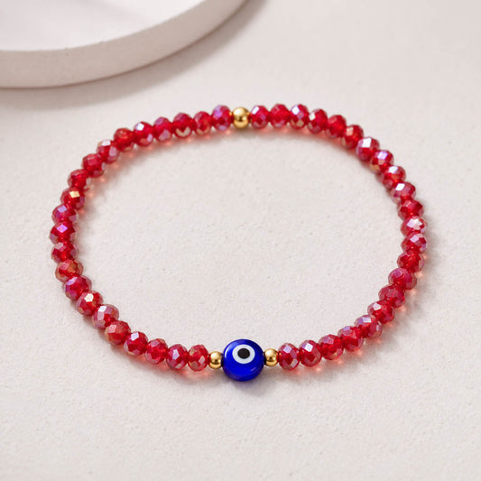 A beaded stretch bracelet made of faceted red beads with a central blue evil eye charm and small gold spacer beads, photographed on a light neutral surface.