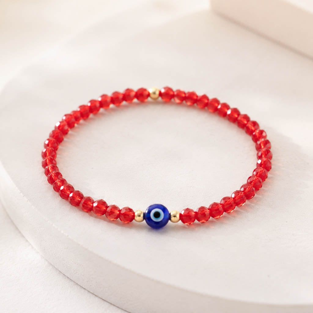A red beaded bracelet with a central blue evil eye charm and small gold spacer beads, displayed on a soft white surface.