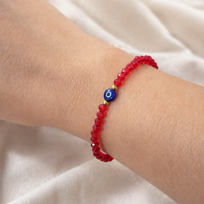 A red beaded bracelet with a blue and white evil eye charm, shown worn on a person's wrist against a soft white fabric background.