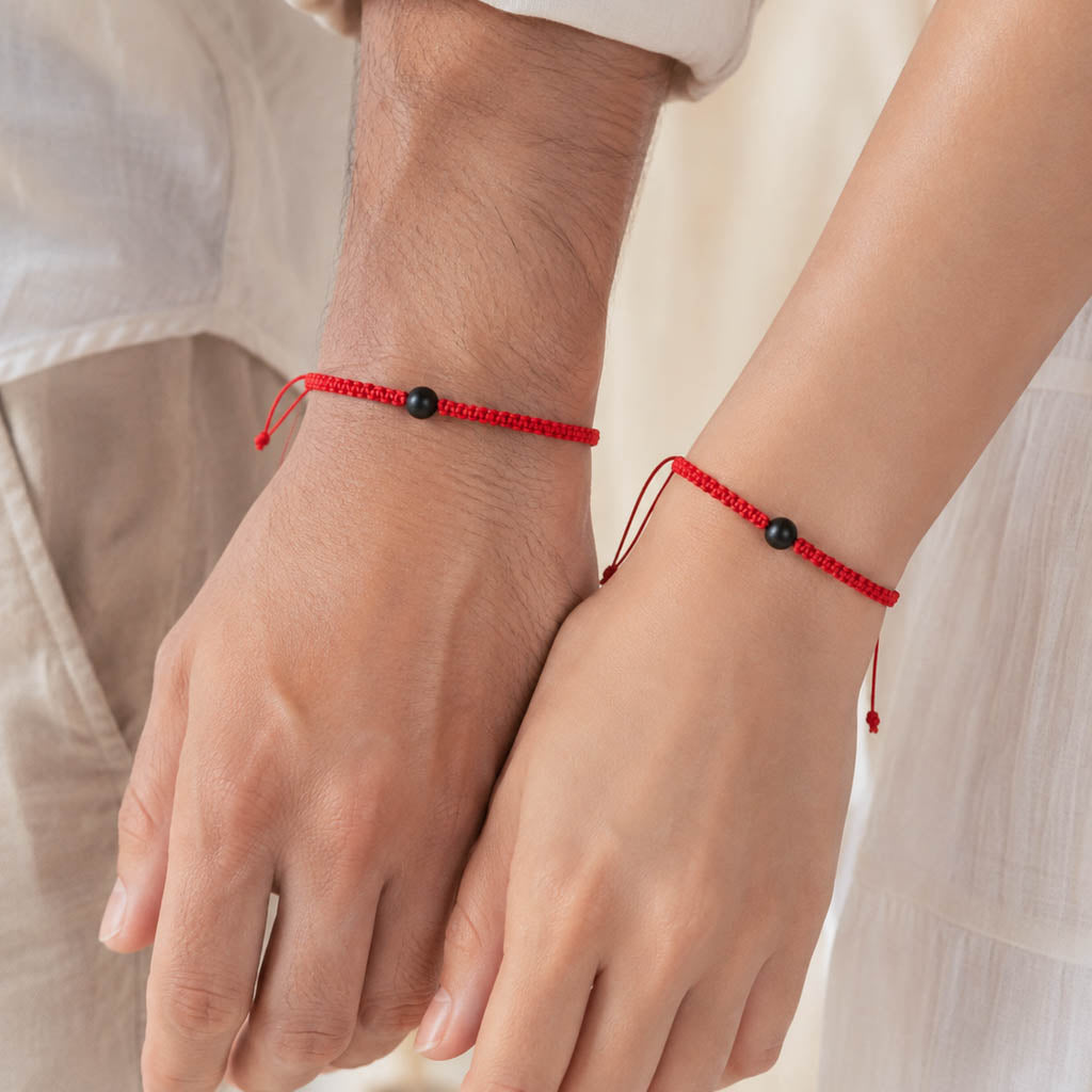Two red cord bracelets with small black beads worn on a person's wrists, shown close up with hands holding hands.