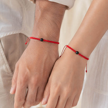 Two red cord bracelets with small black beads worn on a person's wrists, shown close up with hands holding hands.