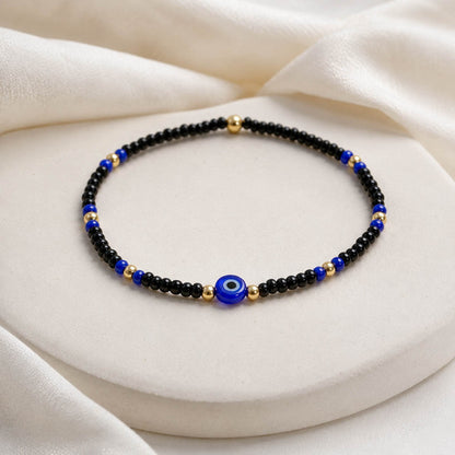 A beaded bracelet with black beads, blue and gold spacer beads, and a central blue evil eye charm, displayed on a cream-colored jewelry stand with draped fabric.