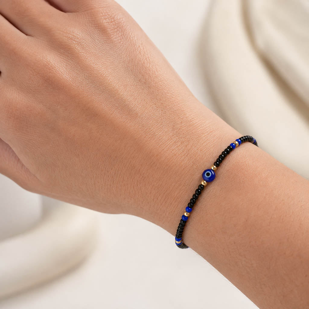 A close-up of a person's wrist wearing a beaded bracelet with black beads, blue evil eye charms, and small gold spacer beads, set against a soft cream-colored background.