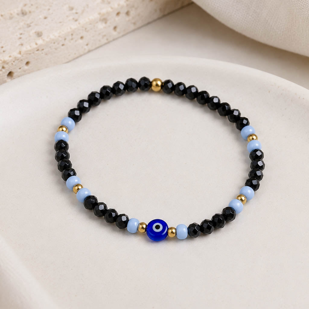 A beaded stretch bracelet featuring faceted black beads, light blue beads, small gold spacer beads, and a central blue evil eye charm, displayed on a light beige ceramic dish.