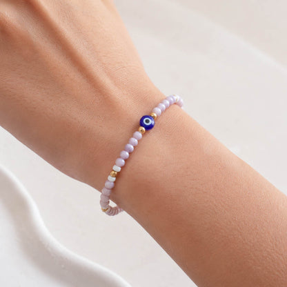 A beaded stretch bracelet with light purple faceted beads, white beads, gold spacer beads, and a blue evil eye charm.