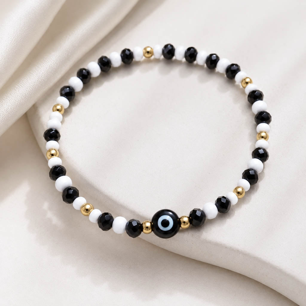 A beaded stretch bracelet featuring alternating black and white beads, accented with small gold spacer beads and a central black evil eye charm with a light blue center, displayed on draped cream satin fabric.