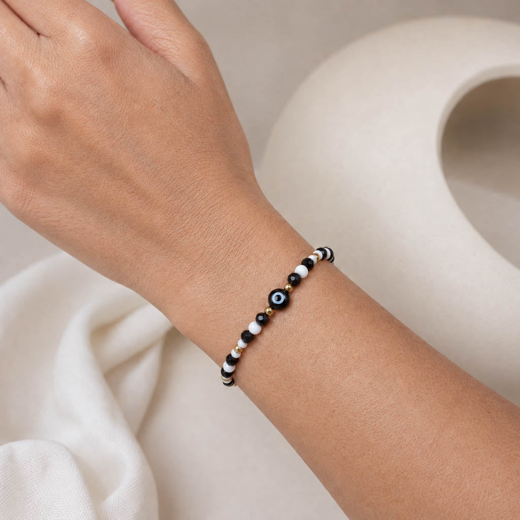 A close-up of a person's wrist wearing a beaded bracelet with black, white, and gold evil eye beads, set against a soft beige background with a white ceramic object.