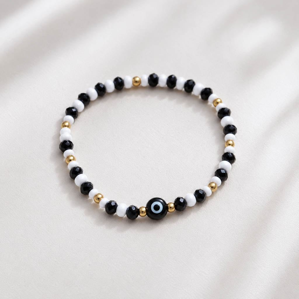 A beaded stretch bracelet featuring alternating black and white beads, accented with small gold spacer beads and a central black evil eye charm with a blue and white center, displayed on a soft, light beige fabric background.