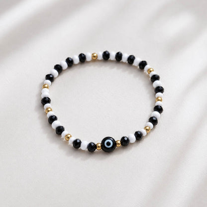 A beaded stretch bracelet featuring alternating black and white beads, accented with small gold spacer beads and a central black evil eye charm with a blue and white center, displayed on a soft, light beige fabric background.