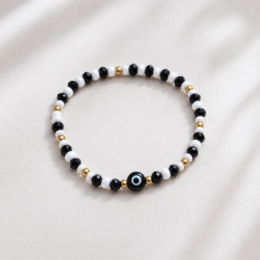 A beaded stretch bracelet featuring alternating black and white beads, accented with small gold spacer beads and a central black evil eye charm with a blue and white center, displayed on a soft, light beige fabric background.