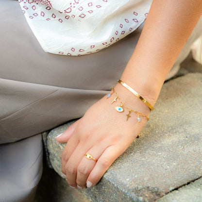 A close-up of a person's hand resting on a stone surface, wearing a gold bracelet stack with a flat band, a delicate chain with small charms, and a matching gold ring.