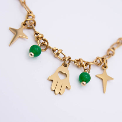 Gold-tone charm necklace featuring a Hamsa hand, two star charms, and green bead accents on a delicate chain, shown against a plain white background.