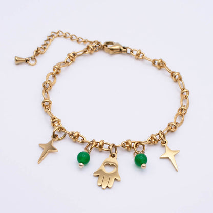 A gold chain bracelet with dangling charms, including two green beads, a Hamsa hand charm, and two star charms, displayed on a clean white background.