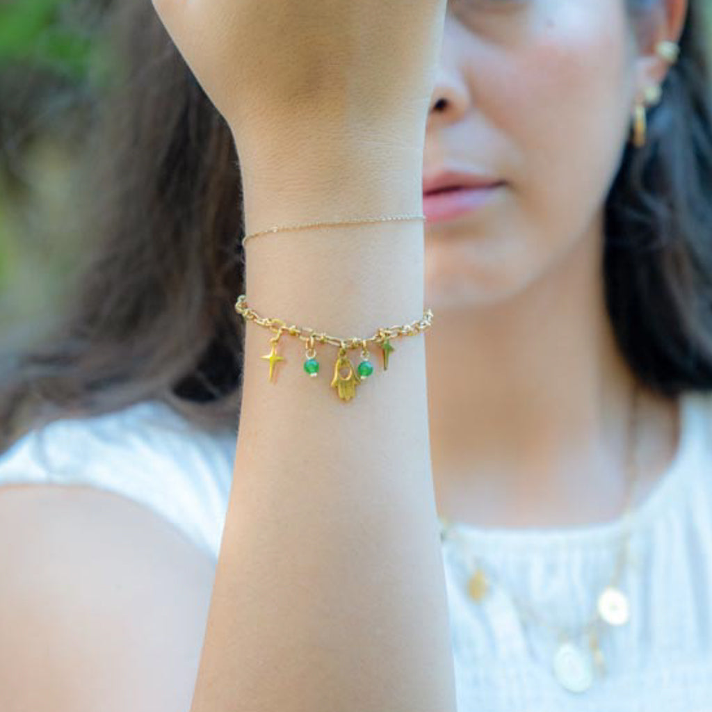 A delicate gold chain bracelet with small hanging charms, including a hamsa hand and green gemstone accents, worn on a woman's wrist in an outdoor setting.