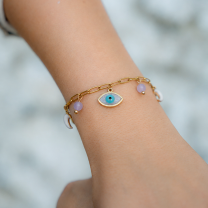 A delicate gold chain bracelet with an evil eye charm and small pink beads, shown worn on a person's wrist against a soft, blurred light gray background.