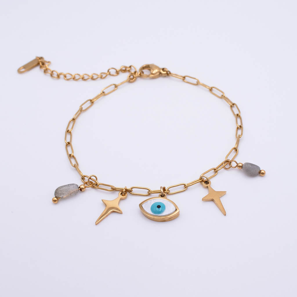A gold chain anklet featuring a central evil eye charm with a blue iris, flanked by two gold star charms, a small gray stone bead, and a larger polished gray stone bead.