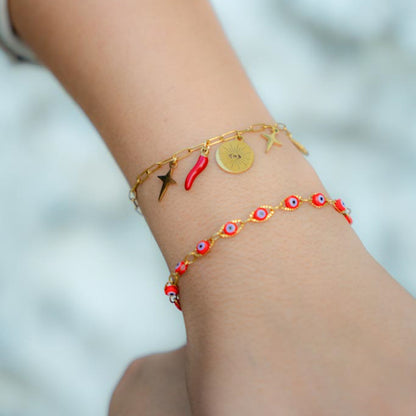 Two delicate gold-tone bracelets worn on a person's wrist. The top bracelet features gold charms including a sun disc, star, and red chili pepper.