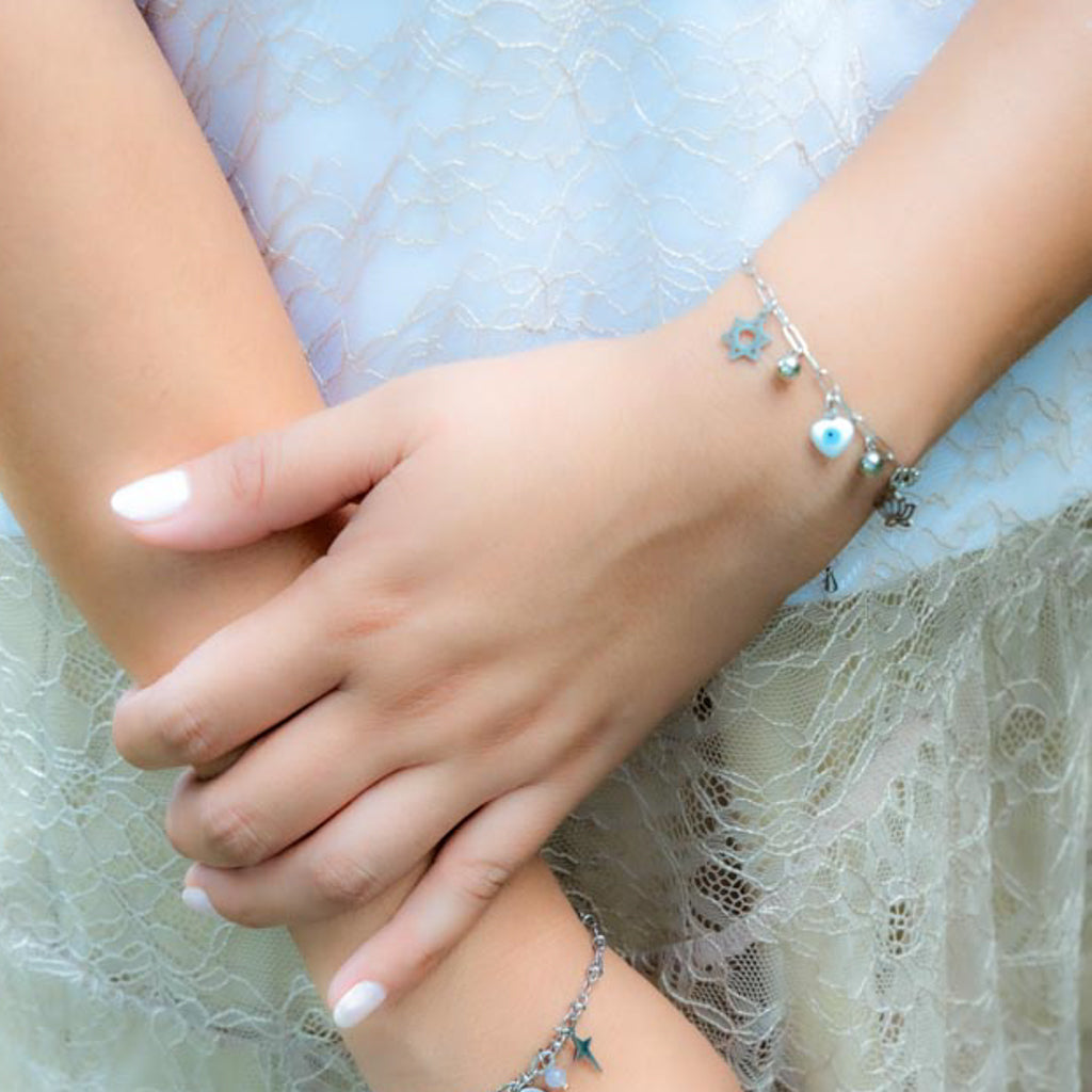 A close-up of a person's hands and arms wearing delicate silver charm bracelets, including a star and evil eye design, against a light blue lace dress.