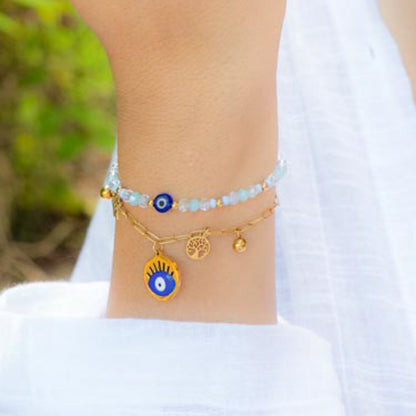 A close-up of a person's wrist wearing two delicate bracelets: a light blue beaded bracelet with a blue evil eye charm, and a gold-tone chain bracelet with a tree of life charm and a blue evil eye charm.