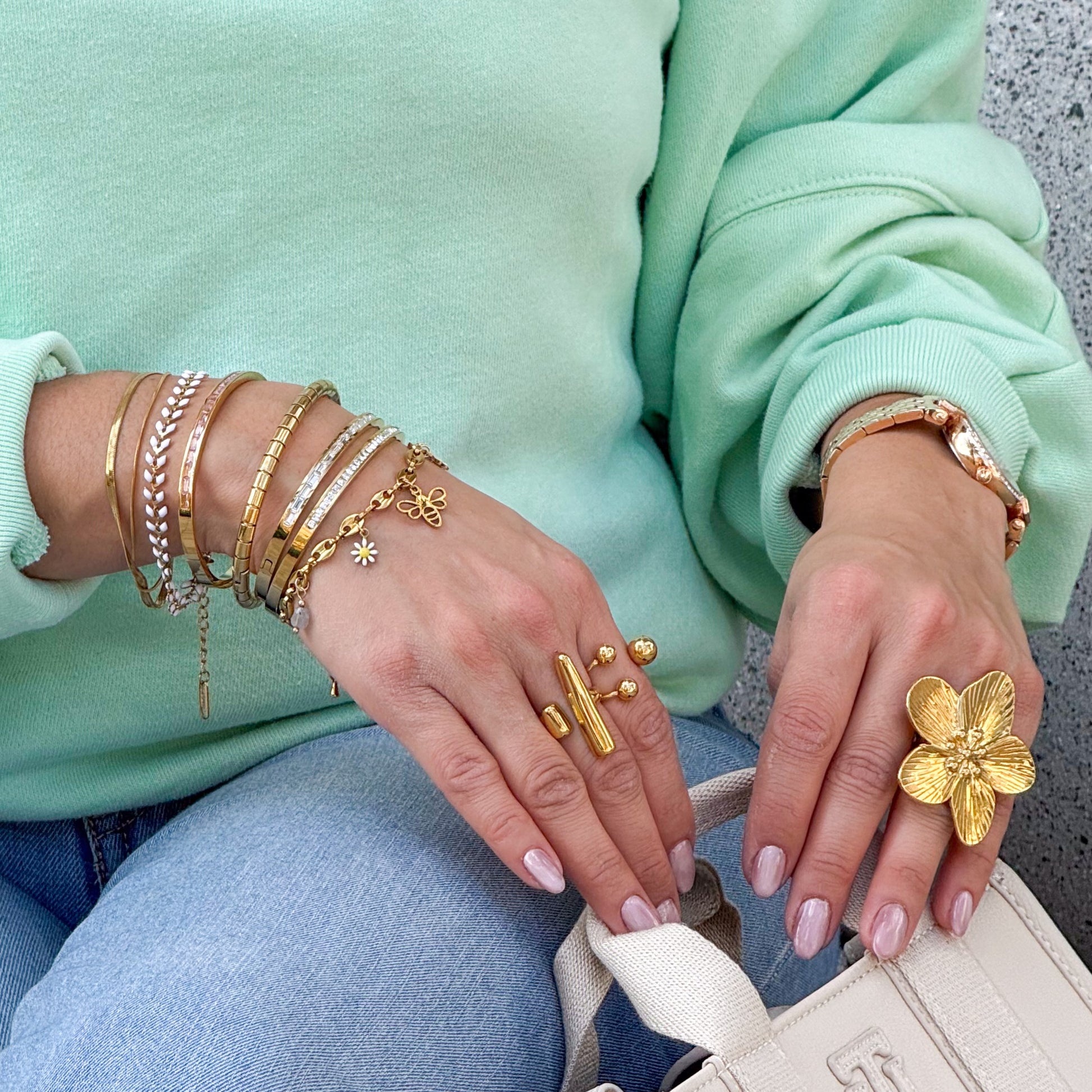 Close-up of hands wearing multiple gold bracelets and rings, with a light green sweater and blue jeans.