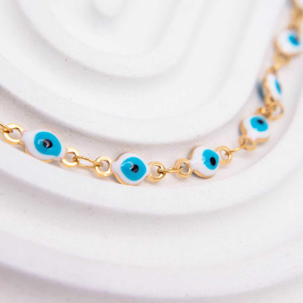 Gold chain necklace with white and blue evil eye beads, displayed on a white textured background.